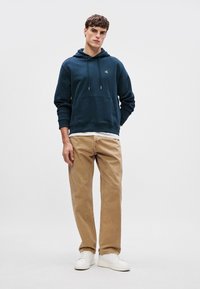 Felpa a cappuccio blu navy con tasca frontale, abbinata a pantaloni beige chiaro e sneakers bianche, in piedi davanti a uno sfondo neutro.