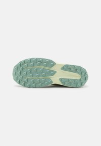 Semelle d'une chaussure de trail légère de couleur claire avec des crampons verts épais en forme de triangles, conçus pour améliorer l'adhérence et la stabilité sur des terrains accidentés.