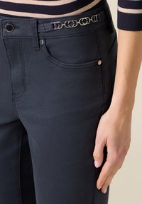 Close-up van een hand rustend bij de heup, gedragen bij donkerblauwe broek met zilveren decoratieve kettingdetail op de tailleband.