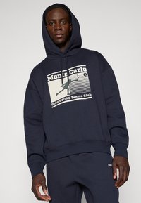 Μπλέ ναυτικού φούτερ με κουκούλα που διαθέτει εκτυπωμένο σχέδιο με "Monte Carlo" και έναν τενίστα. Οι ριβέ τελειώματα στους καρπούς και το στρίφωμα προσφέρουν δομή.