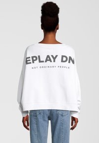 Mujer con el cabello recogido en un moño, vestida con un sudadera blanca de gran tamaño que lleva el texto "REPLAY DN" y "NOT ORDINARY PEOPLE" en la parte trasera, y unos jeans azules.