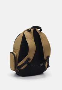 Mochila marrón y negra con correas de hombro acolchadas, hebilla en el pecho y bolsillo lateral, mostrada desde atrás sobre un fondo blanco.