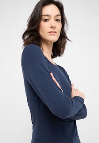 Marineblaues Langarmoberteil aus weichem Baumwollstoff; mit einem runden Halsausschnitt und tailliertem Schnitt. Das Model verschränkt die Arme und zeigt die Ärmellänge.