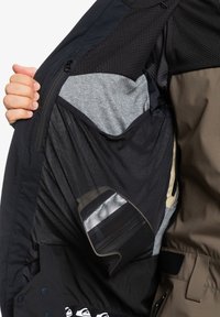 Interior de chaqueta aislante con un bolsillo de malla, un forro gris y tejido negro visible con un compartimento transparente para almacenamiento.