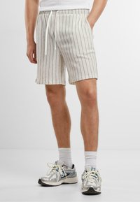 Pantalones cortos gris claro con rayas verticales, cinturilla elástica con cordón, hasta la rodilla, tejido texturizado, con bolsillos laterales, combinados con zapatillas.