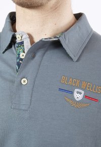 Polo gris avec un col intérieur à motif, deux boutons et du texte brodé. Présente des rayures d'accent bleu et rouge et une couture décorative.
