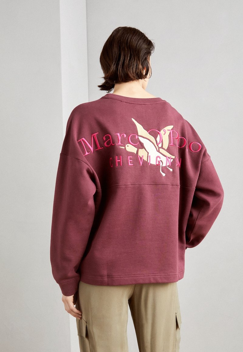 Personne portant un sweat-shirt ample bordeaux avec un texte rose "Marc O'Polo Chevignon" et un graphisme blanc d'oiseau dans le dos, associé à un pantalon beige.