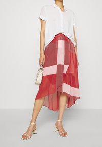 Jupe fluide à carreaux rouges et roses avec un ourlet asymétrique, associée à une chemise blanche boutonnée et des sandales à talons beige. Tient un petit sac.