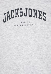 Siva majica kratkih rukava s crnim tiskanim tekstom "JACK&JONES" koji se proteže na vrhu, ispod "EST. 90" i "WORLDWIDE" manjim fontom. Mekana tekstura.