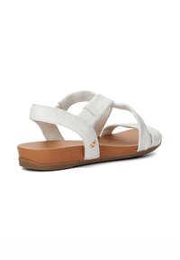 White leather sandals with cross straps, a flat brown outsole, and a small gold accent detail. Smooth texture and casual design.