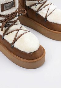 Botas de ante marrón con forro de piel de oveja blanca, cordones decorativos marrones y suela gruesa color marrón claro. Cuenta con un parche de logo en el lado.