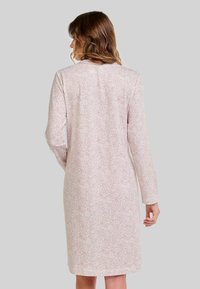 Mujer con cabello castaño ondulado, vestida con un vestido de manga larga, hasta la rodilla, de color rosa claro con un pequeño patrón de puntos, vista desde la espalda.