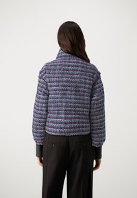 Korte jas in een multicolored tweed stof met een hoge kraag en gathered mouwen, gecombineerd met zwarte op maat gemaakte broeken.