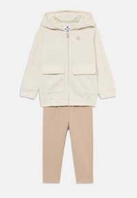 Krämfärgad zip-up hoodie med två framfickor och en logga; ihop med beige leggings. Mjuk tyg, avslappnad passform och enkel design.
