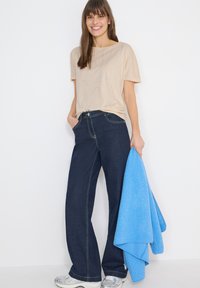 Femme souriante aux longs cheveux bruns portant un haut beige à manches courtes et un jean large foncé, tenant une veste polaire bleu vif dans la main droite.