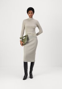 Lichtbeige geribbelde trui met hoge hals, gecombineerd met een bijpassende geribbelde midi-jurk; model houdt een groene leren tas vast. Zwarte kniehoge laarzen.