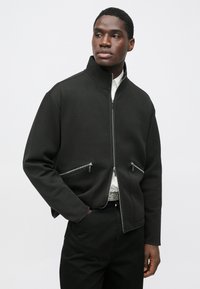 Schwarze Reißverschlussjacke mit hohem Kragen, ausgestattet mit zwei vorderen Reißverschlusstaschen und einem glatten, strukturierten Stoff. Kontrastierende Details an den Nähten.