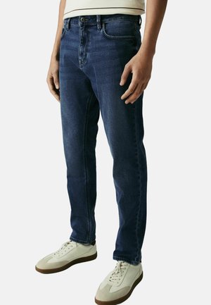 Dark blue denim jeans with a slim fit, featuring neutral stitching, five pockets, and a front button closure. Worn with white sneakers.