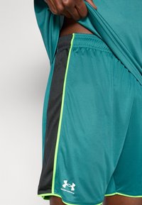 Tirkīzzilas sporta šorti ar melnām sānu daļām, neona zaļiem akcentiem un elastīgu jostasvietu. Apakšējā labajā pusē atrodas Under Armour logo.