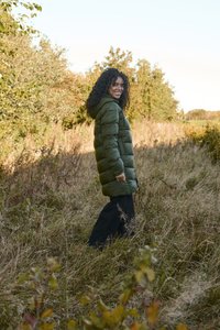 Padded green coat with a hood, featuring a quilted design and long sleeves, paired with black pants, in a grassy outdoor setting.