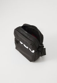 Bolso cruzado de nylon negro con una superficie texturizada, cierre de cremallera y un gran logo blanco en la parte frontal. Tiene una correa ajustable y un bolsillo interior.