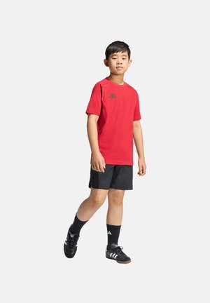 Ragazzo che cammina indossando una maglietta sportiva rossa, pantaloncini neri, calze Adidas nere e scarpe sportive Adidas nere su uno sfondo chiaro uniforme.