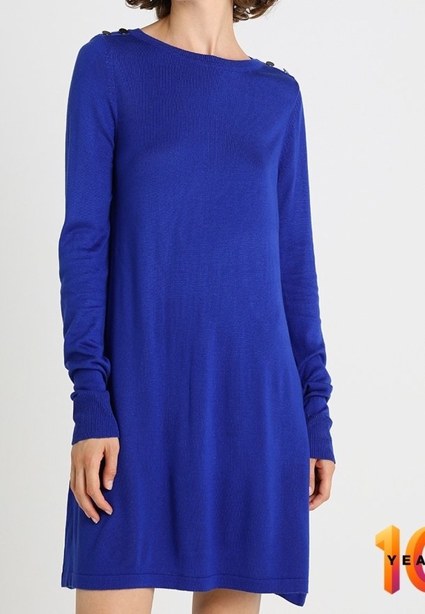 Frau, die ein langärmliges, knielanges, königsblaues Strickkleid mit Knöpfen an den Schulterähnen trägt und vor einem schlichten hellen Hintergrund steht.
