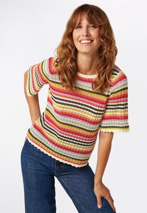 Meerachtige gestreepte trui met een zigzagpatroon, korte mouwen, gegolfde randen en een lichte gebreide textuur. Gecombineerd met een blauwe spijkerbroek.