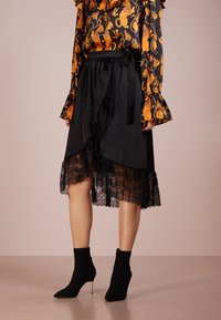 Jupe noire midi avec un design en couches agrémenté de dentelle, associée à une finition satinée, et une ceinture à nouer assortie à la taille.