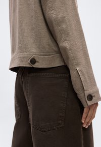 Posnetek od blizu osebe, ki nosi bež majico z dolgimi rokavi z zapetimi gumbi na manšetah in temno rjave hlače z vidnim zadnjim žepom.