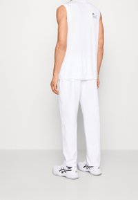 Man i vit ärmlös skjorta, vita byxor och vita gympaskor står vänd bortåt mot en enfärgad bakgrund.