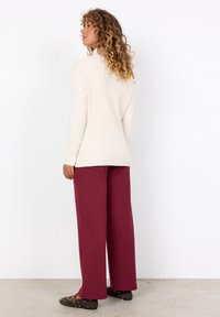Maglione a coste crema, maniche lunghe, abbinato a pantaloni larghi bordeaux. Ballerine marroni con dettagli dorati, sfondo neutro.