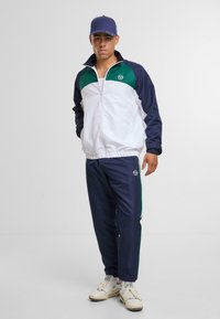 Tuta in bianco, blu navy e verde; giacca con zip e colletto alto con logo; pantaloni con cintura elastica; sneakers texture.