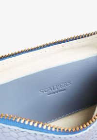 Interior de bolsa de cuero azul claro con una cremallera dorada y forro de tela beige. "SCALPERS HECHO EN ESPAÑA" embossado en el lateral.