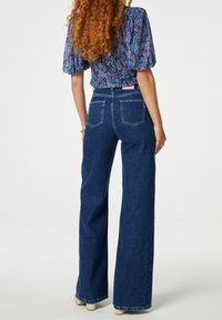 Jeans in denim a gamba larga di colore blu scuro con cinque tasche, cuciture robuste e una leggera texture. Il retro presenta una piccola etichetta del marchio in rosa.