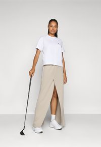 Valge lühike t-särk logoga, beež jagatud seelik ja valged tossud; hoiab golfikeppi; sile kangas, igapäevane spordirõivaste disain.