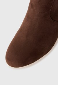 Prim-plan al vârfului rotunjit al unei cizme de gleznă din suede maro, cu cusături vizibile, pe un fundal alb.