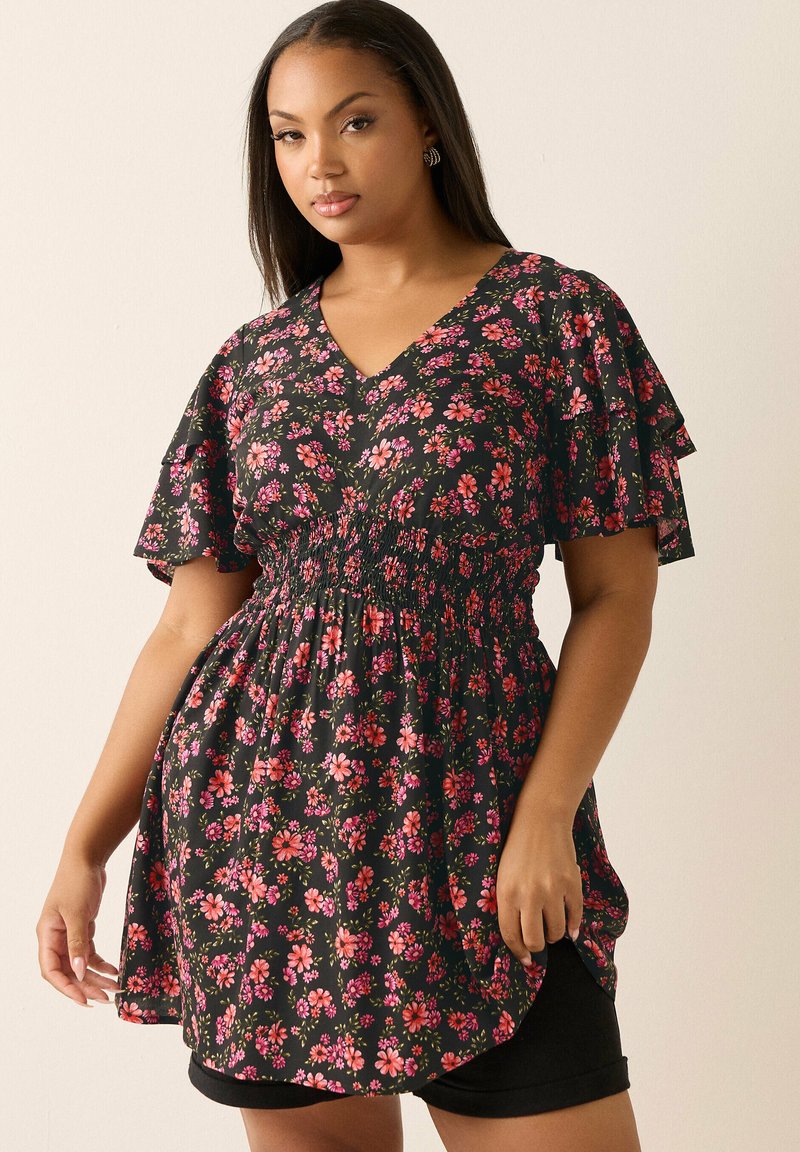 Femme portant une robe noire à fleurs roses et rouges, avec des manches flottantes et une taille smockée, debout devant un fond clair uni.