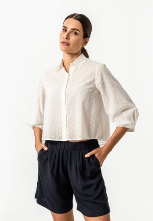 Woman with dark hair in a ponytail wearing a white textured button-up blouse with 3/4 sleeves and black elastic waist shorts, hands in pockets.
