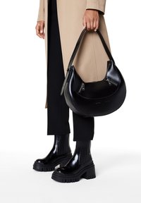 Sac en cuir noir à bandoulière avec un corps arrondi, des accents de fermeture éclair contrastants et une texture lisse. Associé à des bottes noires à talons épais.
