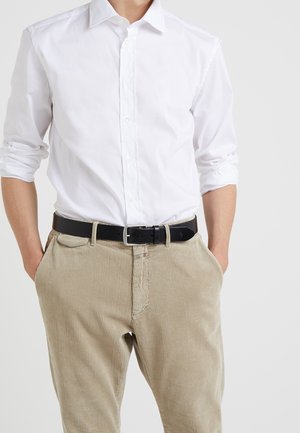 Mężczyzna w białej koszuli zapinanej na guziki z podwiniętymi rękawami oraz beżowych sztruksowych spodniach z czarnym paskiem, ręce w kieszeniach.