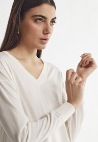 Beige V-Ausschnitt Pullover mit langen Ärmeln und gerippten Bündchen. Weiche Textur, lockere Passform und dezente Nahtdetails am Halsausschnitt.