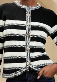Čierno-biely pruhovaný kardigán s okrúhlym výstrihom, zlatými gombíkmi a textúrovanou pleteninou. Obsahuje kontrastný houndstooth vzor na okrajoch.