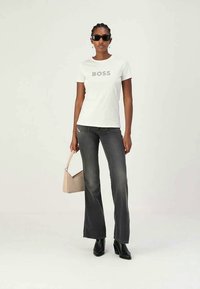 Mujer con gafas de sol, camiseta blanca de "BOSS", jeans de campana negros desgastados, botas negras y sosteniendo un bolso beige frente a un fondo liso.