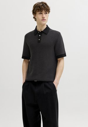 Fiatal férfi göndör hajjal, fekete, texturált, rövid ujjú polóinget és bő, fekete nadrágot visel, kezét a zsebében tartja, balra néz.