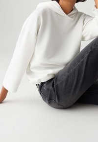Personne portant un sweat à capuche blanc texturé avec cordon de serrage et un jean gris foncé, assise sur une surface gris clair dans une posture détendue.