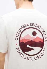 Λευκό βαμβακερό t-shirt με κυκλικό γραφικό σχεδιασμό που απεικονίζει βουνά σε ροζ αποχρώσεις, έναν ποταμό και κείμενο: «Columbia Sportswear Portland, Oregon.»