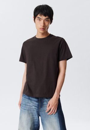 Jeune homme portant un t-shirt à manches courtes de couleur brun foncé uni et un jean bleu délavé, se tenant devant un fond gris clair uni.
