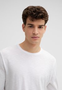 Junger Mann mit lockigem braunem Haar, der ein schlichtes weißes T-Shirt mit Rundhalsausschnitt trägt, vor einem neutralen hellgrauen Hintergrund.