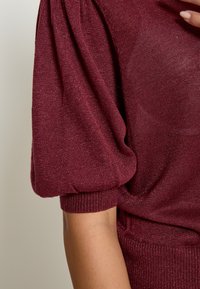 Bordeaux gebreide trui met textuur, puffed korte mouwen en geribbelde onderkant. Subtiele glans voegt visuele interesse toe.